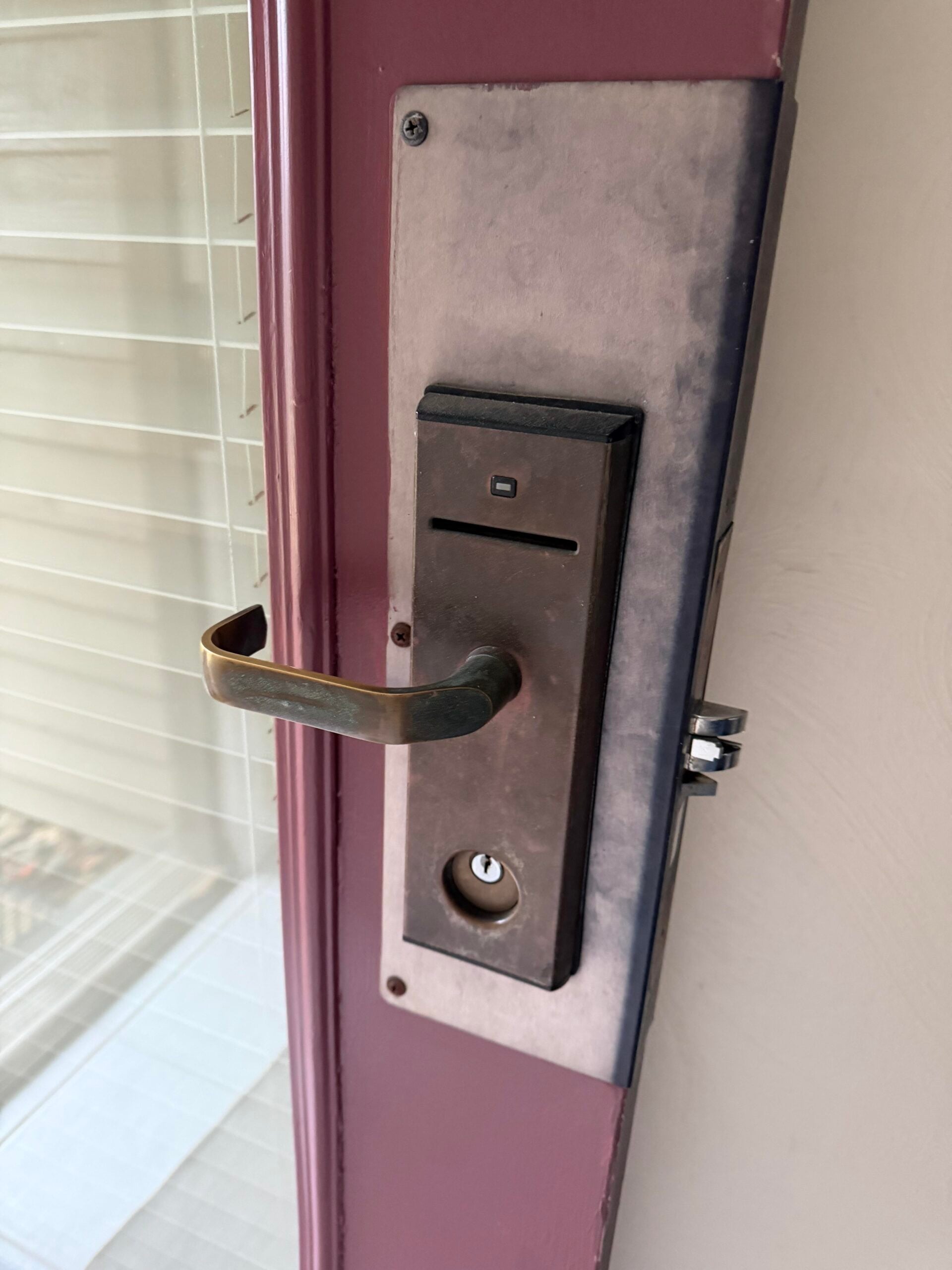 Close-up of a door with a metallic electronic lock and a brass handle. The door is maroon with a glass panel, and window blinds are visible in the background.