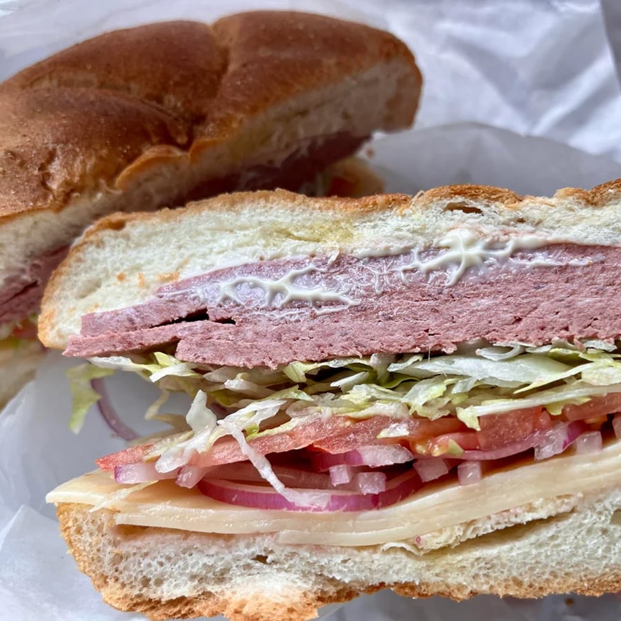 A close-up of a sandwich cut in half, showing layers of lettuce, tomato, red onion, cheese, sliced deli meat, and mayonnaise on a round bread roll. One half is placed in front of the other.