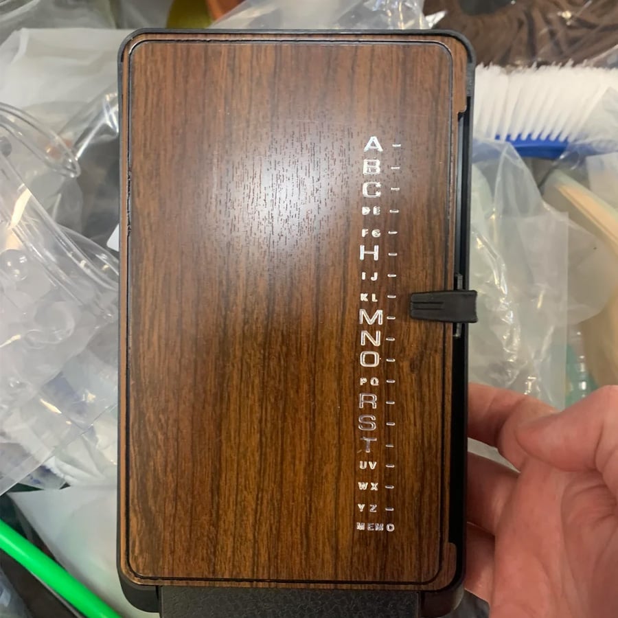 A hand holds a vintage brown plastic device with a faux wood finish and an A-Z list on the side, likely an old address or phone number organizer.
