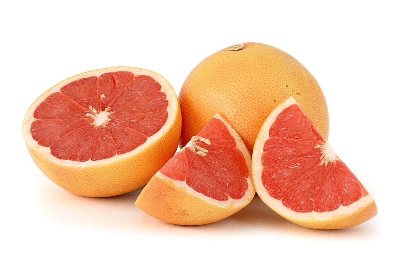 A whole grapefruit, a halved grapefruit showing pink flesh, and two grapefruit wedges are arranged on a white background.