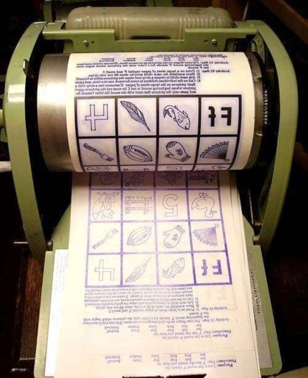 A close-up of a vintage mimeograph machine printing sheets with blue ink. The paper shows various symbols, numbers, and animal illustrations arranged in a grid.