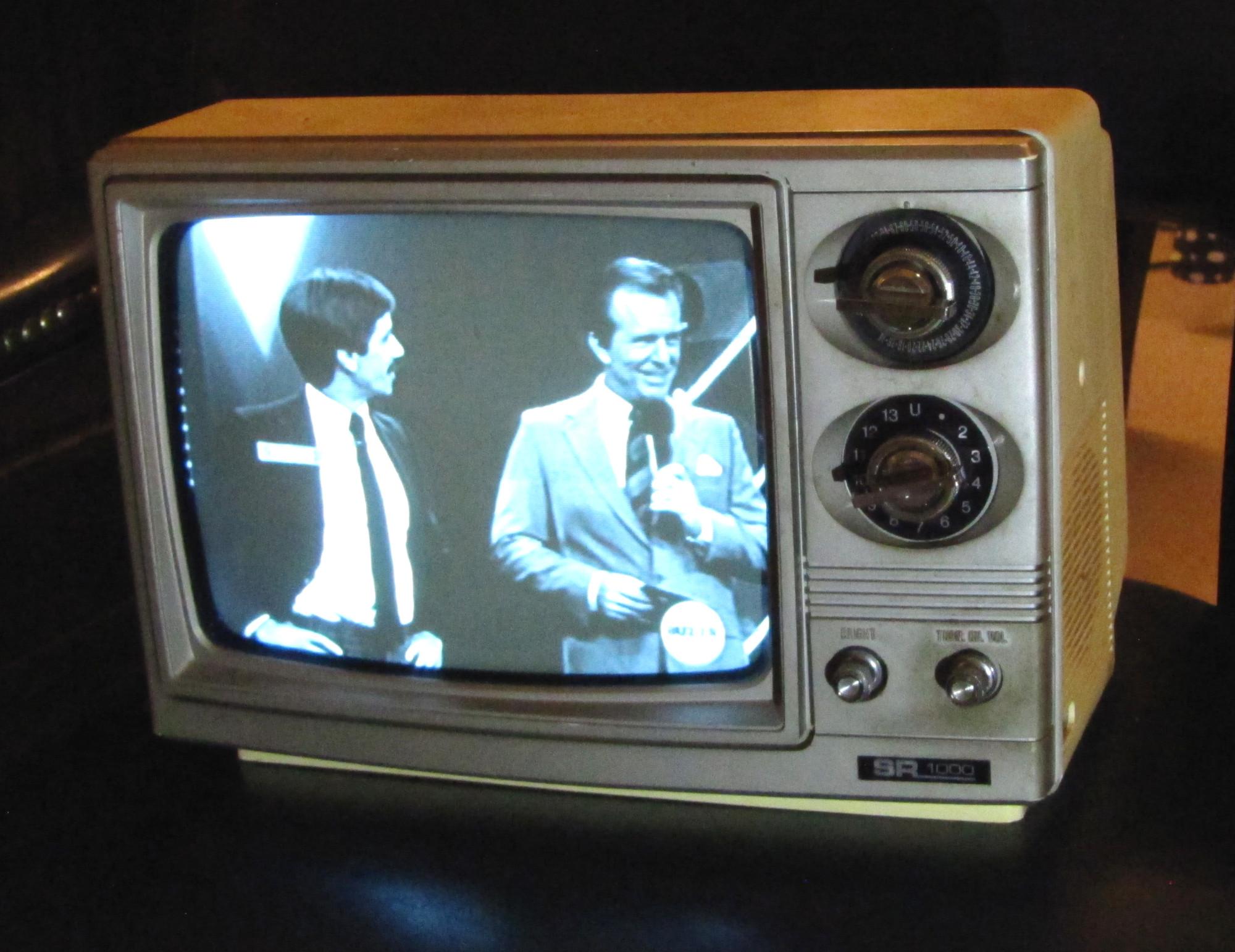 A black-and-white TV displays two men in suits speaking. The TV is vintage, with dials for tuning and volume on the right, and the brand label “SR-1000” visible at the bottom.