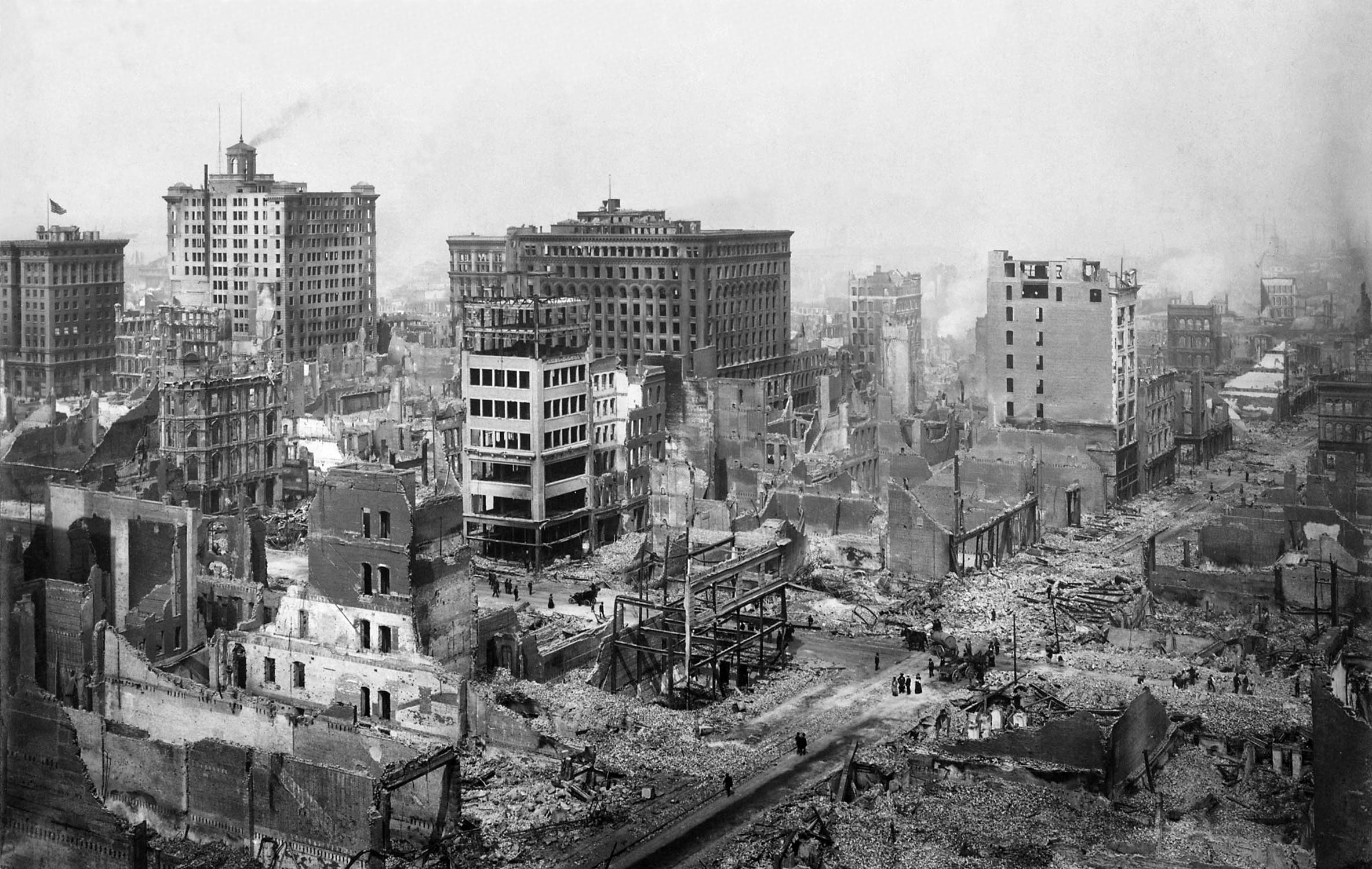 A black and white photo shows a city devastated by disaster, with partially destroyed buildings, rubble-strewn streets, and smoke or haze in the background. A few people walk among the ruins.
