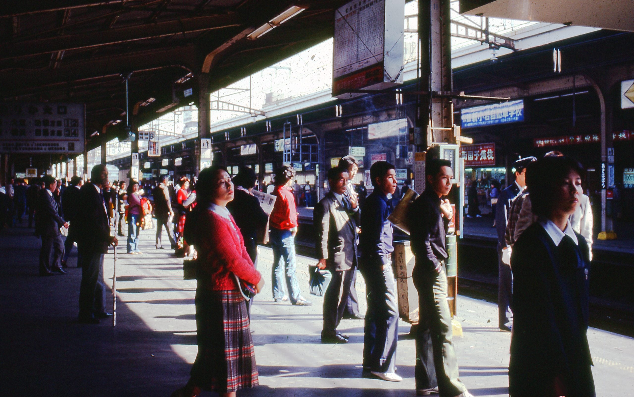 People wait on a busy train platform under a large roof, with sunlight casting shadows. Some look down the track, while others walk or stand in groups. Signs and advertisements hang above the platform.