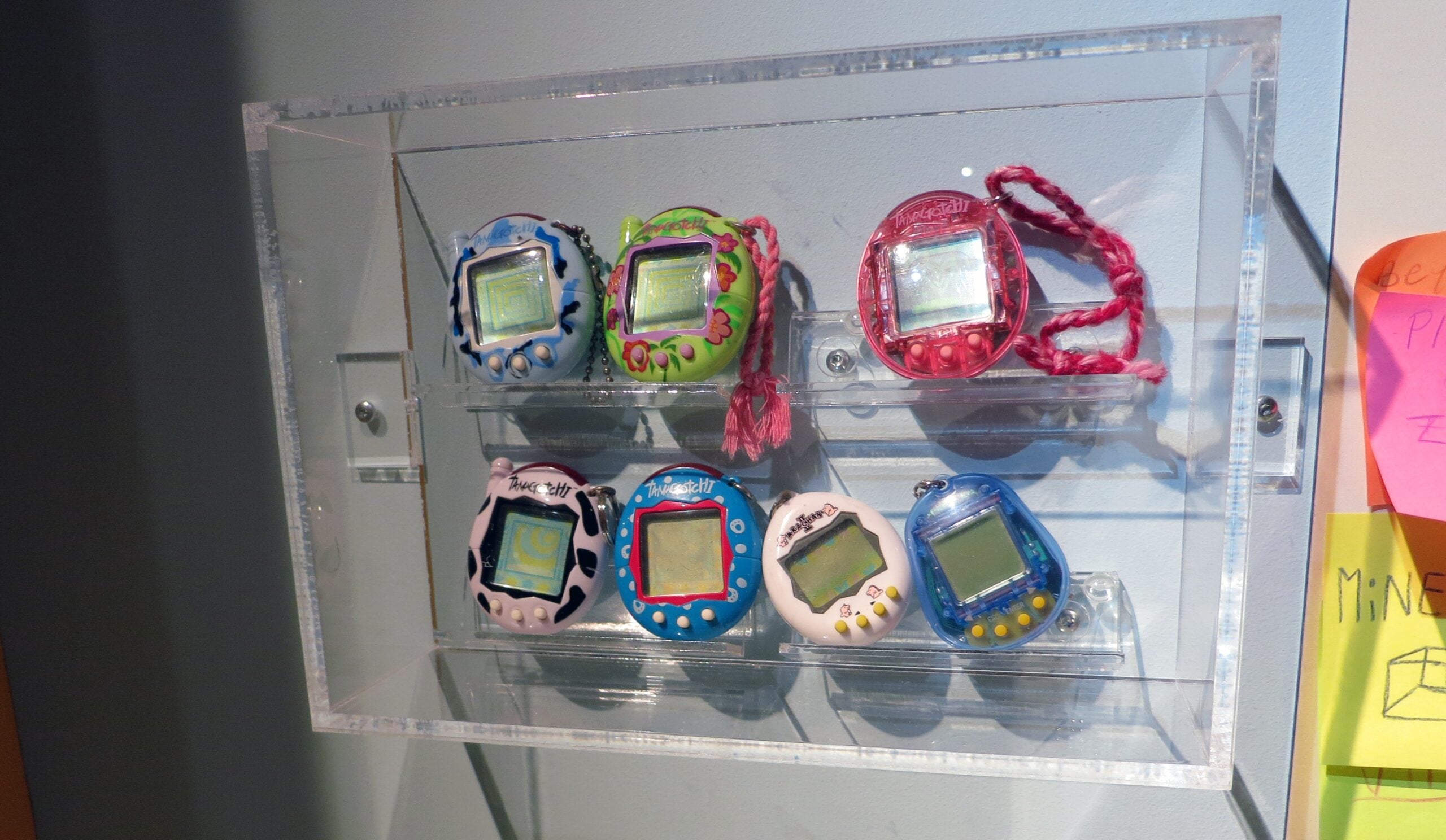 A clear display case mounted on a wall holds seven colorful Tamagotchi digital pets arranged on two shelves. Bright sticky notes are partially visible on the right edge.