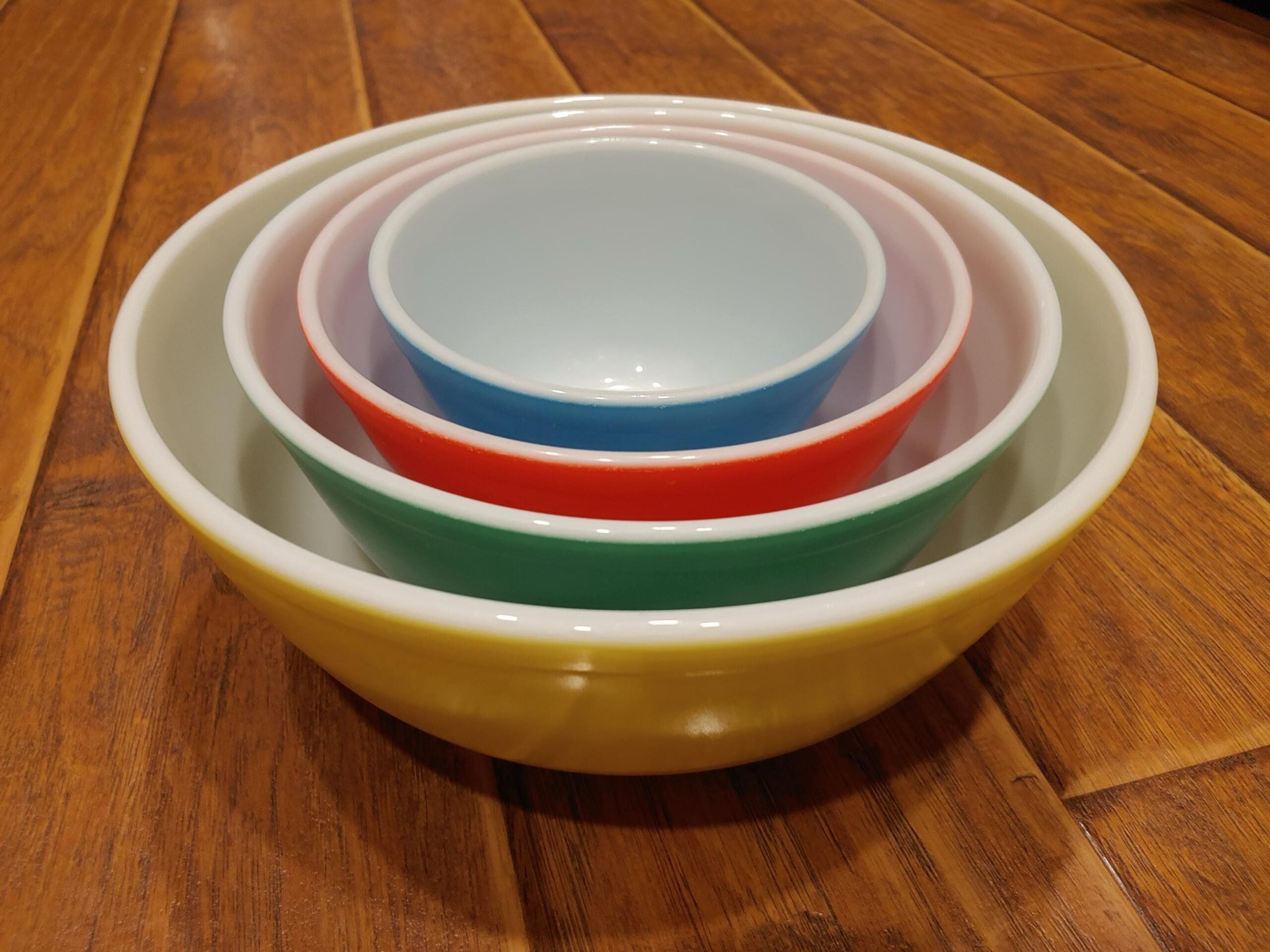 Four colorful mixing bowls—yellow, green, red, and blue—nest inside each other on a wooden surface. The bowls decrease in size from outermost to innermost, creating a concentric arrangement.