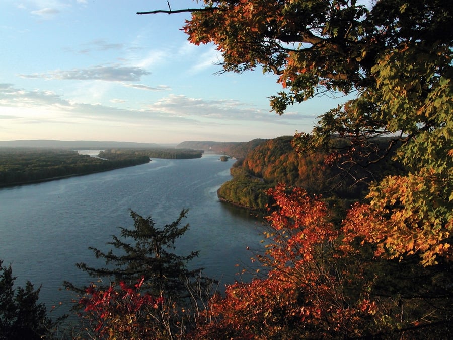 A wide river curves through a landscape of green and orange trees under a partly cloudy sky, with autumn leaves framing the scene in the foreground.