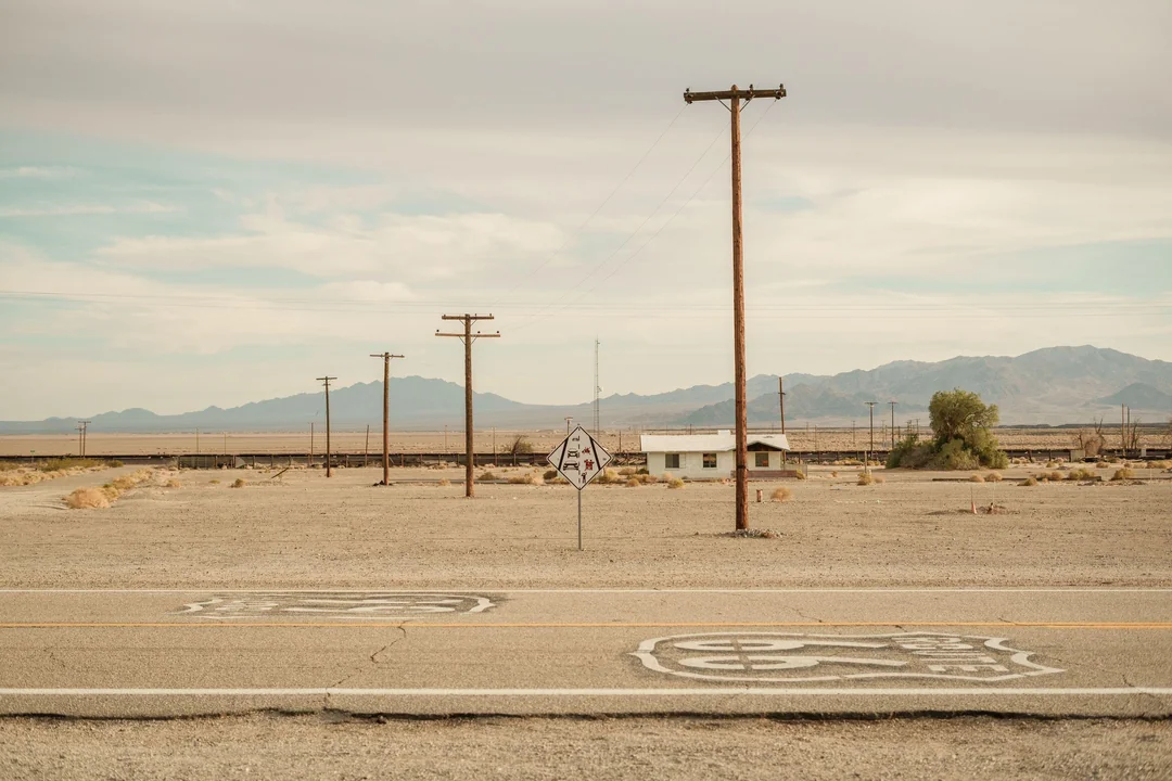 A deserted landscape with a small building, utility poles, a faded road sign, and Route 66 symbols painted on the road; mountains and cloudy sky in the background.