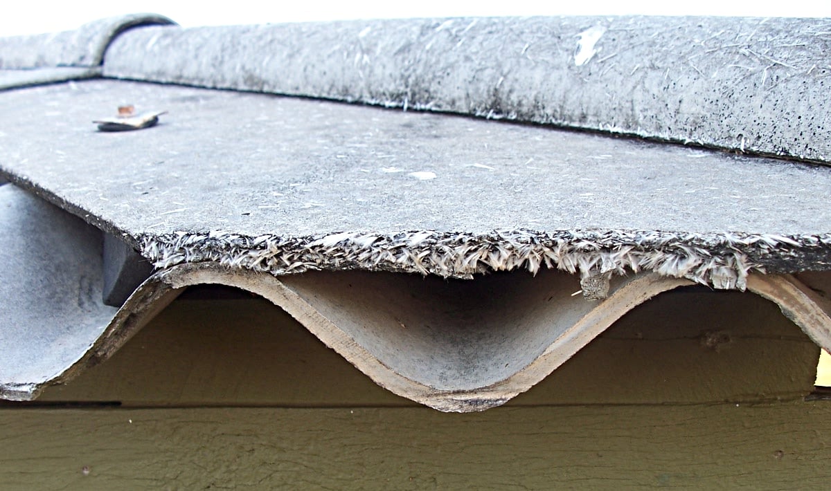 A close-up of a weathered, corrugated asbestos roof panel showing frayed edges, mounted on a building with a green wall underneath.