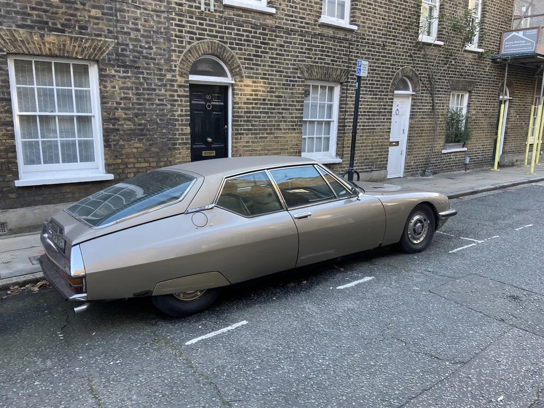 A vintage silver car is parked on a quiet street in front of brick houses with white-trimmed windows and doors. The scene appears calm and residential.