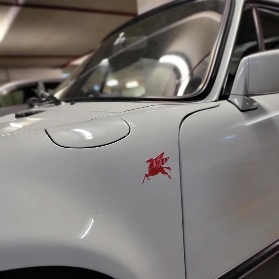 White classic car with a red Pegasus decal on the front fender, parked indoors. The windshield and part of the hood are visible, with blurred background elements.