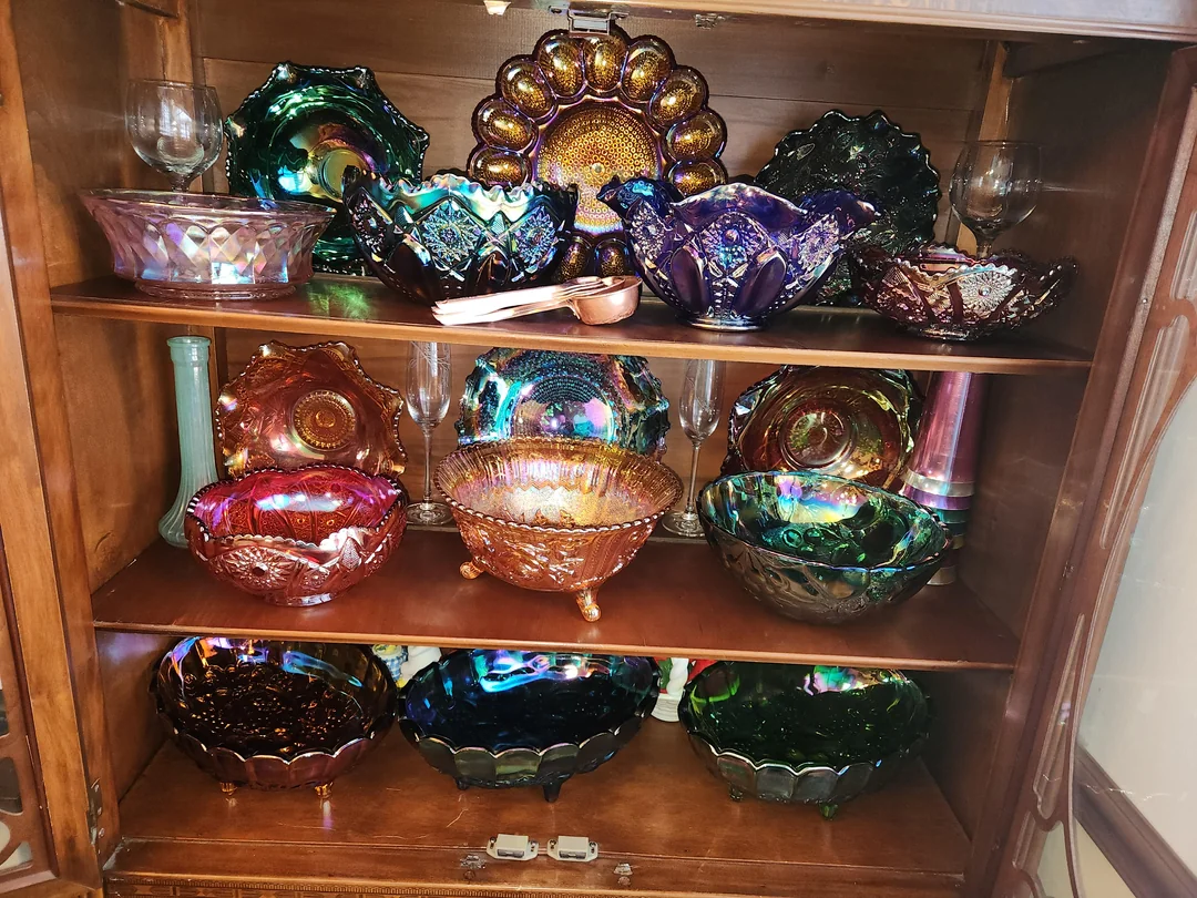 A wooden cabinet with glass doors displays colorful, iridescent carnival glass bowls, plates, and goblets on three shelves. The glassware shines with vibrant hues of purple, green, gold, and blue.