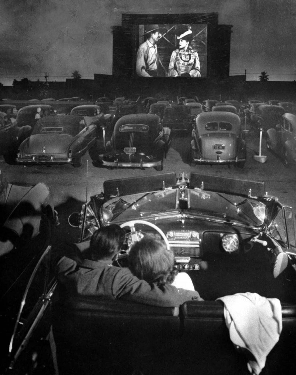 A black-and-white photo of people sitting in classic cars at a drive-in movie theater at night, watching a film projected on a large outdoor screen.