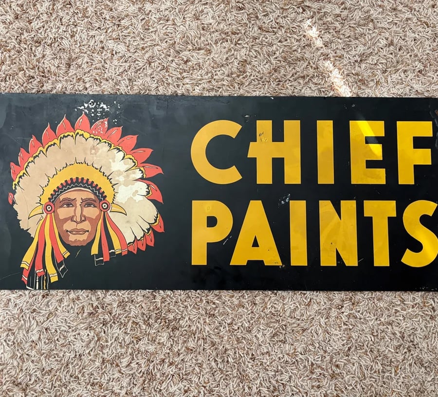 A vintage sign on a carpet reads “CHIEF PAINTS” in yellow letters, with an illustrated Native American chief in a feathered headdress on the left side.