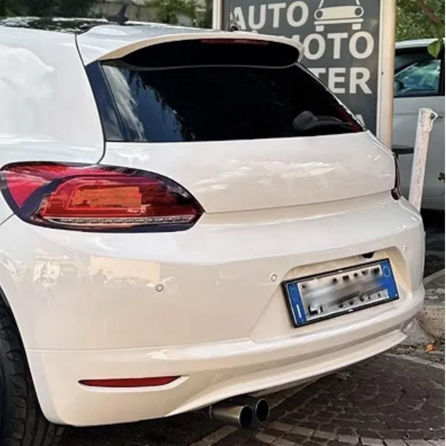 A white hatchback car with tinted rear windows is parked on a street near a sign that partially reads “AUTO MOTO CENTER.” The license plate is blurred.
