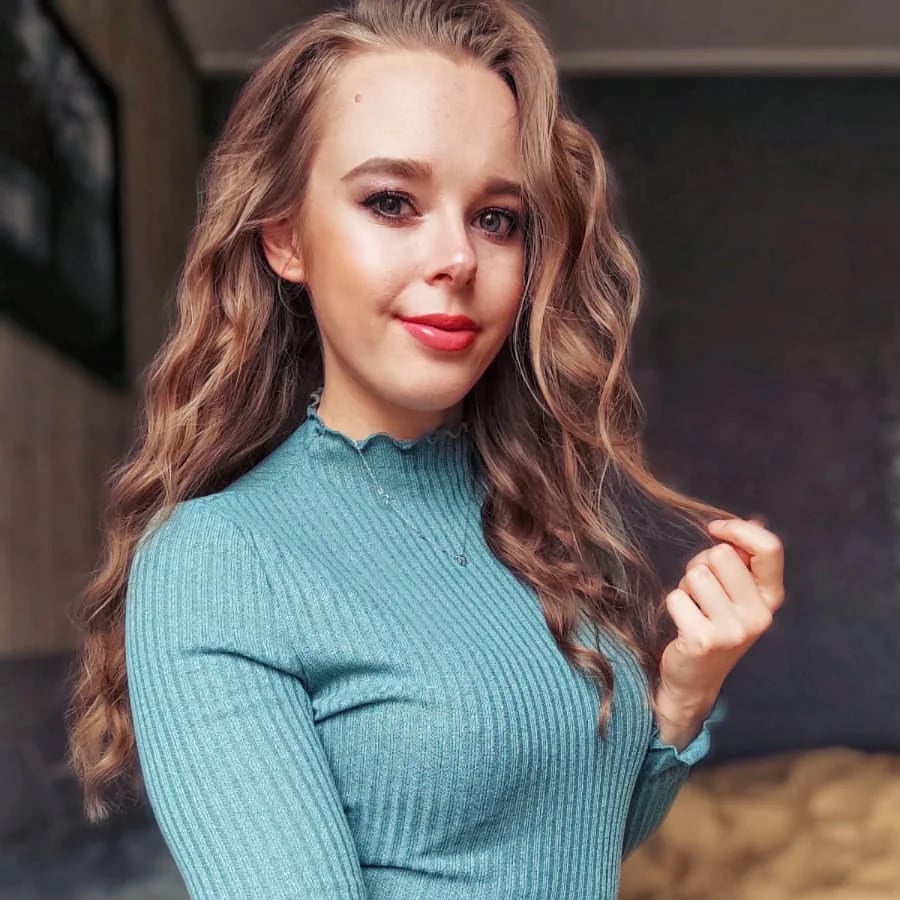 A woman with long, wavy light brown hair wears a ribbed teal top, smiling slightly while holding a strand of her hair. The background is indoors with neutral-colored walls.