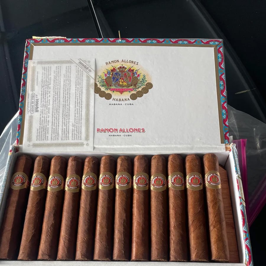 An open box of Ramon Allones cigars from Havana, Cuba, showing thirteen brown cigars neatly lined up inside with decorative labels and a colorful border on the box.