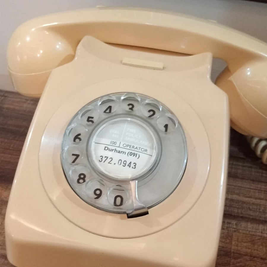 A beige rotary dial telephone sits on a wooden surface. The dial shows numbers 0 to 9, and the center label lists the phone number “372-0943” and the location “Durham (091)”.