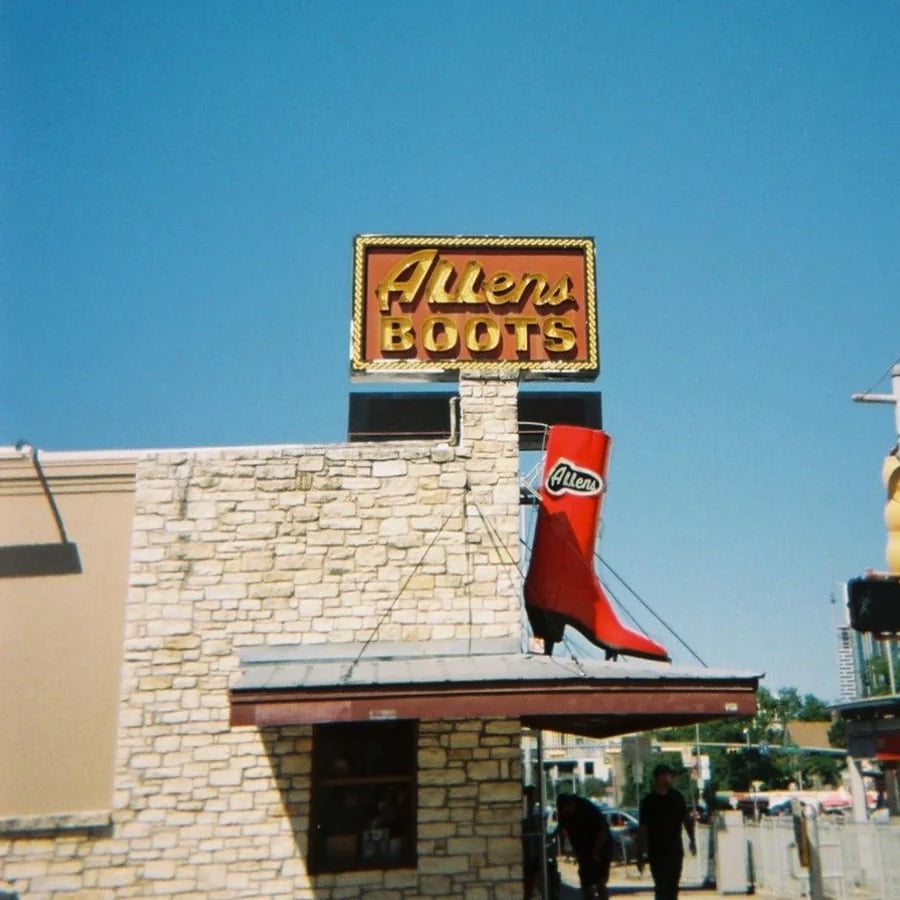A stone building with a large red boot sign and a marquee reading “Allen’s Boots” against a clear blue sky. Two people stand near the entrance.
