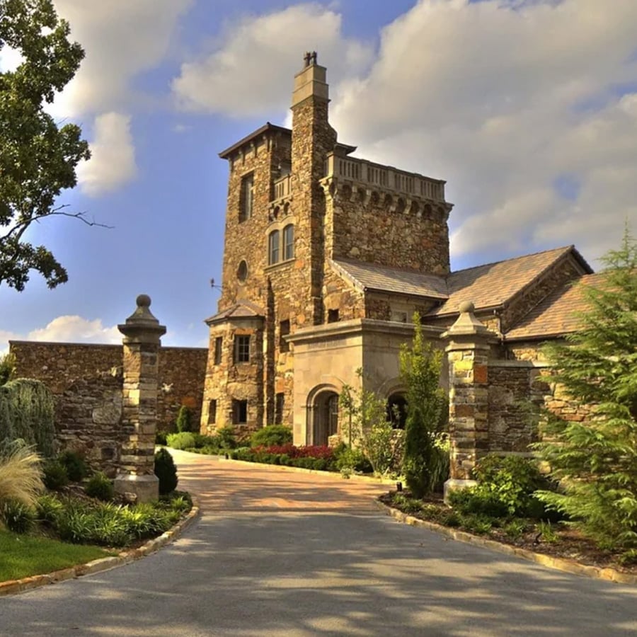 A large stone mansion with a central tower sits behind an iron gate, surrounded by lush greenery and trees under a blue sky with scattered clouds.