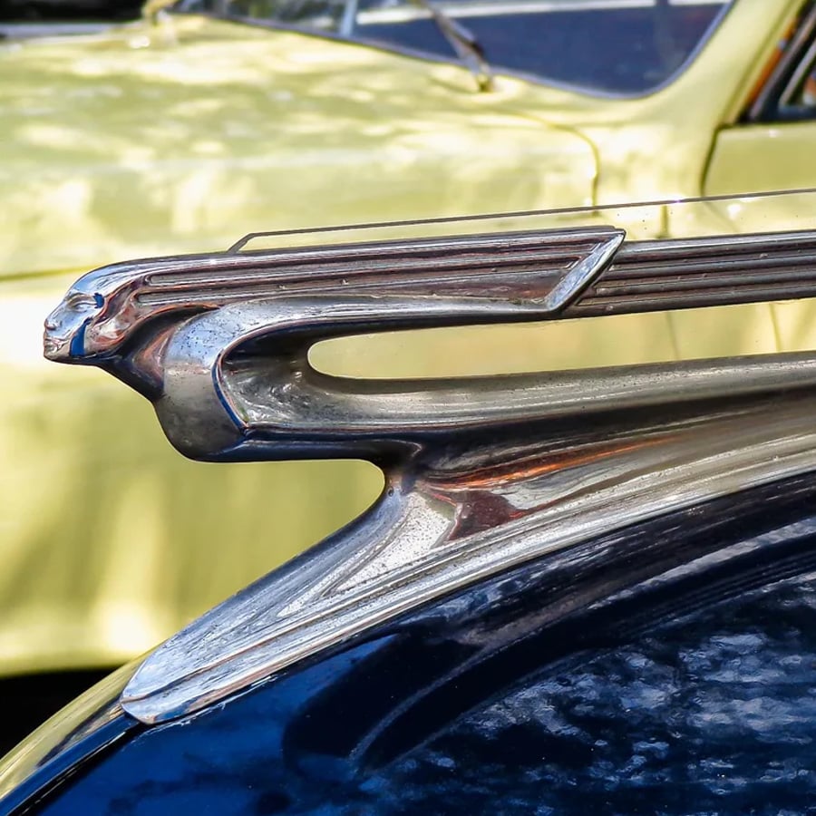 Close-up of a vintage car hood ornament shaped like a streamlined figure, with a yellow car blurred in the background.