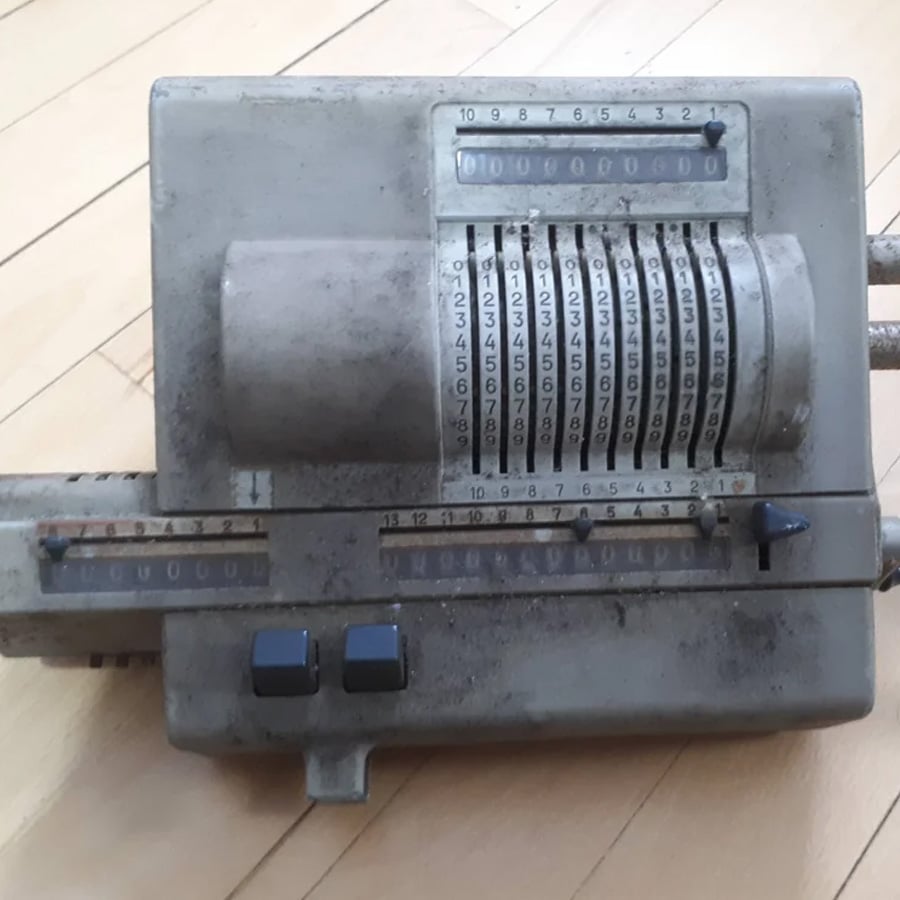 A vintage mechanical calculator with numbered rotating dials and levers sits on a wooden floor. The device appears worn and dusty, showing signs of age and use.