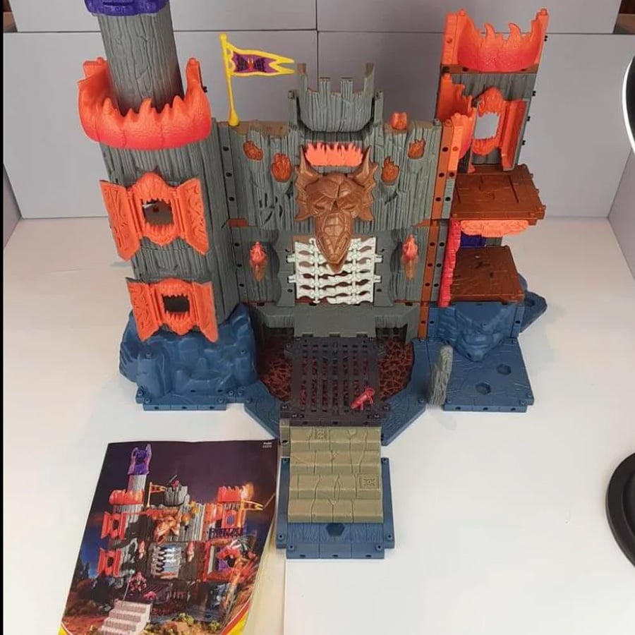 Toy castle playset with gray and red fortress, towers, a yellow flag, and an instruction booklet in front. The castle has rocky blue base and raised platforms on each side.