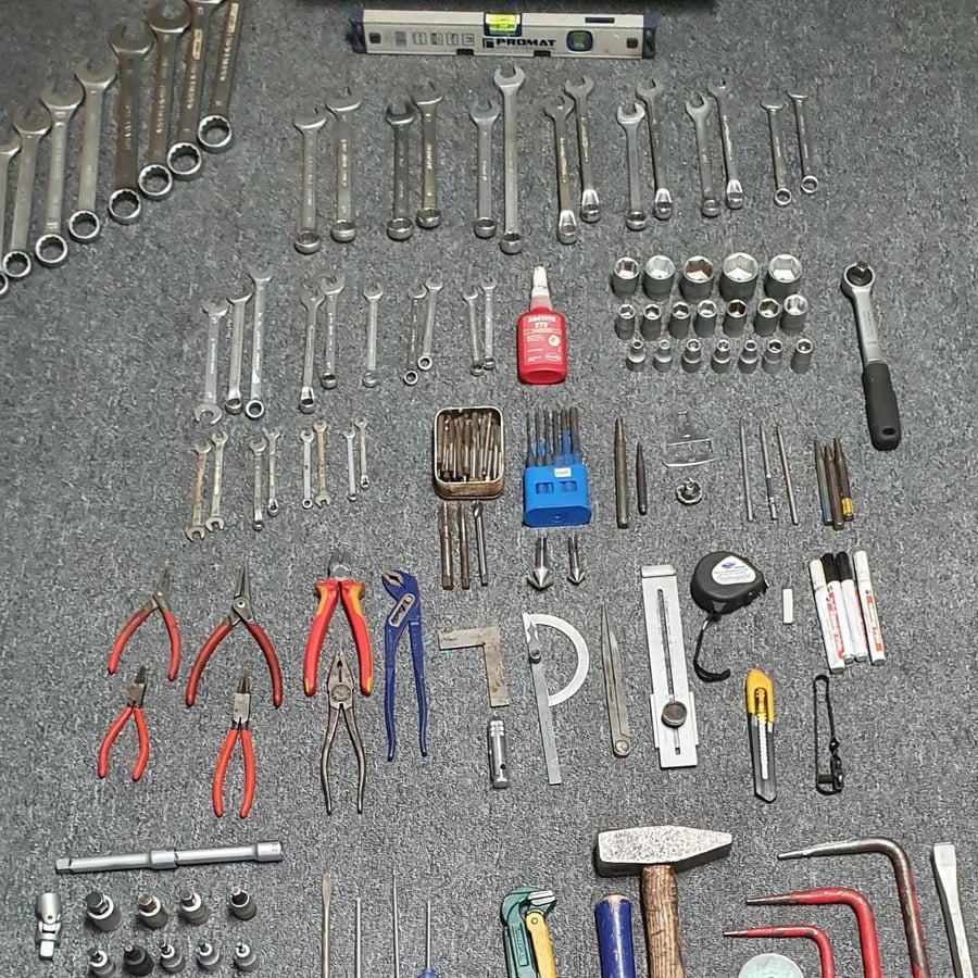 Assorted tools including wrenches, sockets, pliers, screwdrivers, a tape measure, calipers, and a hammer neatly arranged on a gray carpet.