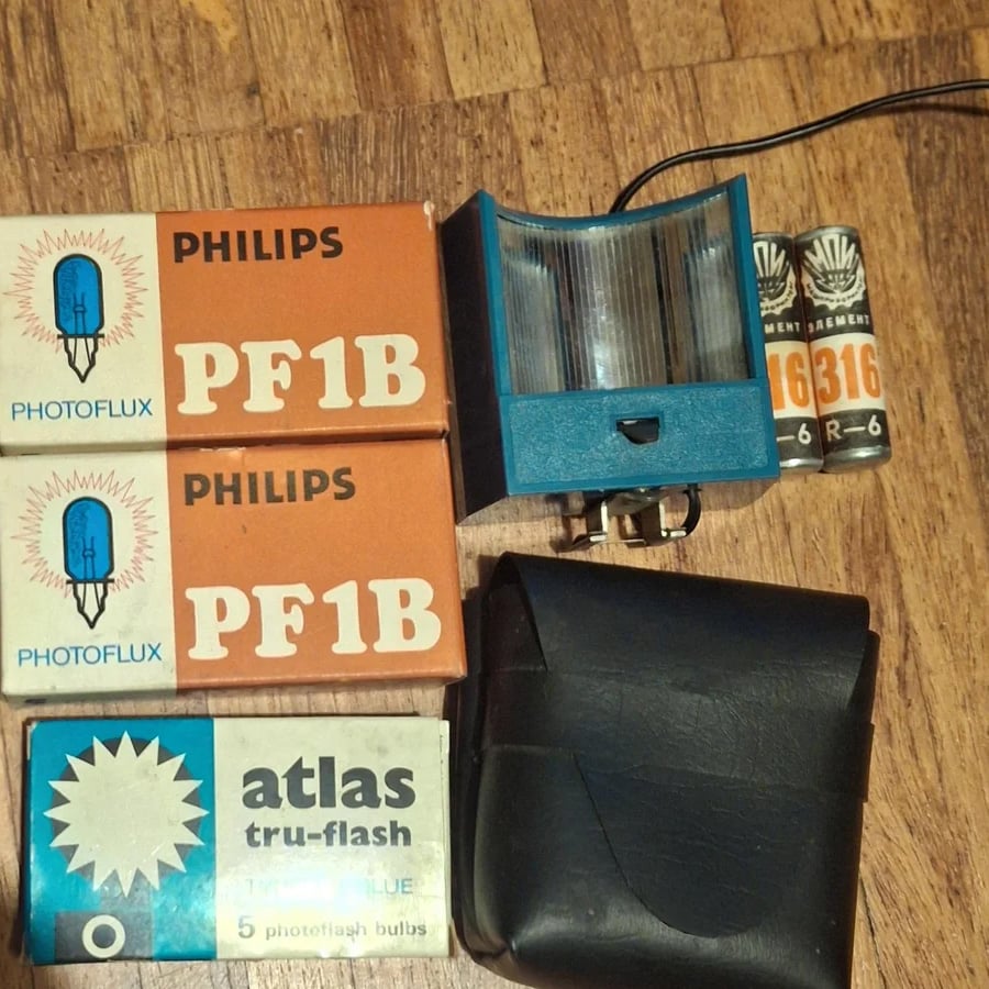 Two Philips PF1B flashbulb boxes, one Atlas tru-flash box, a blue flash unit, a black case, and two Eveready AA batteries are arranged on a wooden floor.
