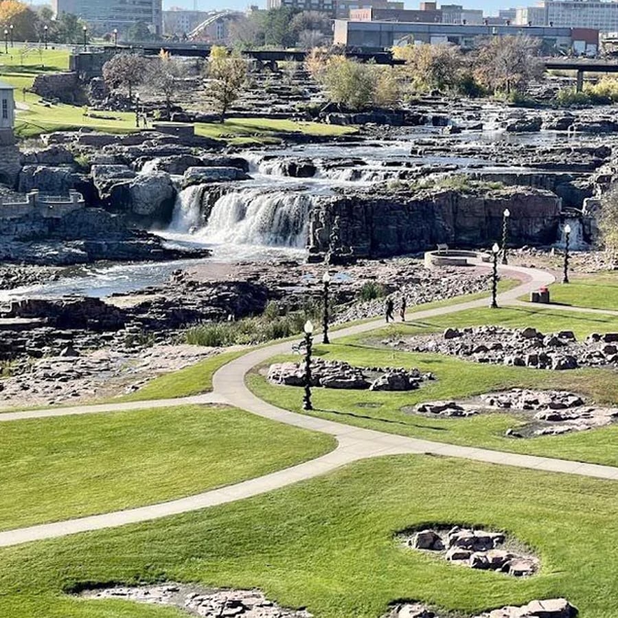 A scenic park features winding paths, green lawns, rock formations, lampposts, and a cascading waterfall in the background, with city buildings visible beyond the trees.