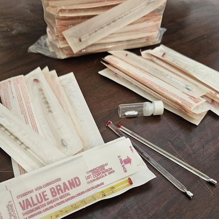 Several glass rectal thermometers in packaging and loose on a wooden surface, along with a small vial and a stack of packaged thermometers in the background.