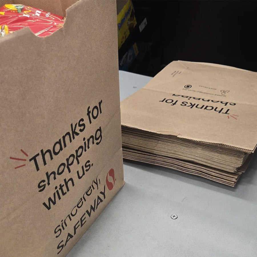 A Safeway paper grocery bag with “Thanks for shopping with us. Sincerely, SAFEWAY” text is upright beside a stack of flat, unused brown bags on a store counter.