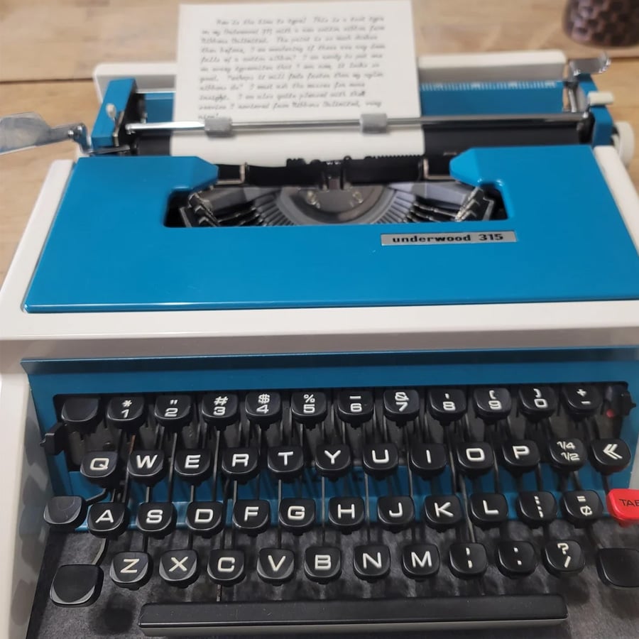 A close-up of a blue Underwood 315 typewriter with a page inserted. The black keys are labeled in white, and the typebars and platen are visible. The typewritten page contains several lines of text.