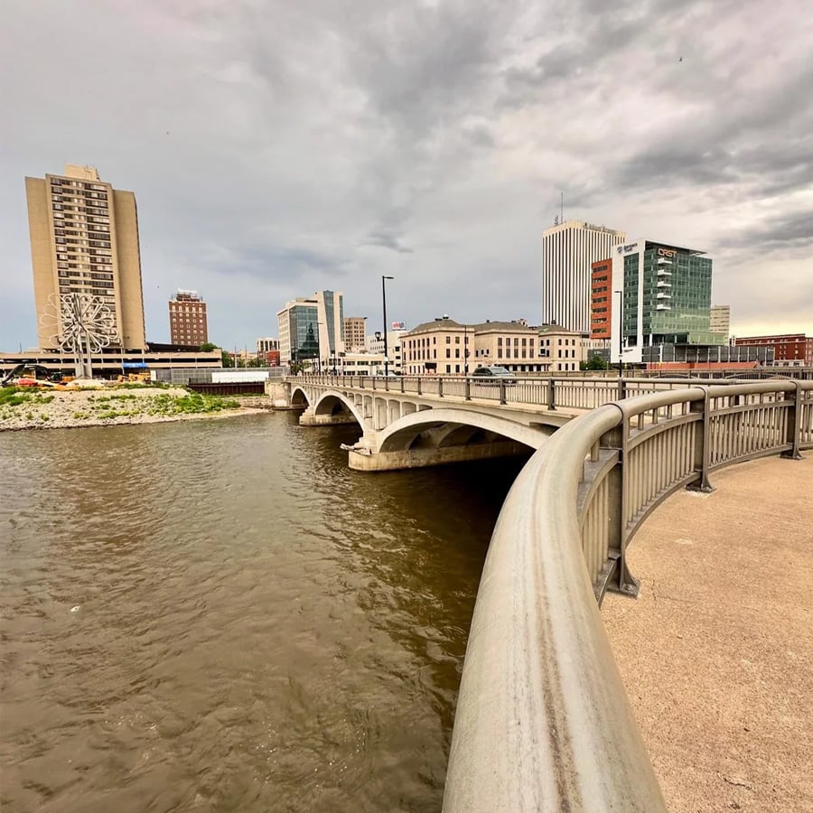 A curved bridge crosses a river with tall city buildings in the background under a cloudy sky. A metal railing lines the bridge and walkway in the foreground.