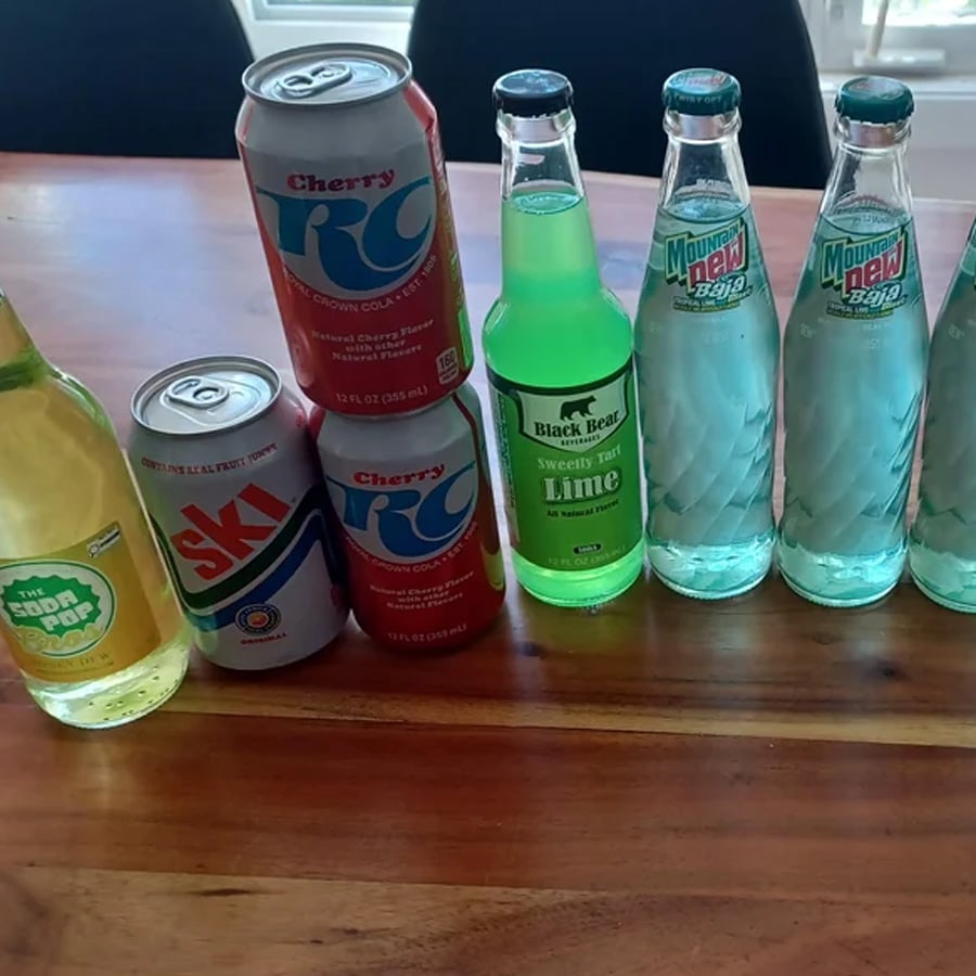 Seven sodas sit on a wooden table, including two Cherry RC cans, a Ski can, an MS Soda Pop bottle, a Black Bear Lime bottle, and two glass bottles of Mountain Dew.