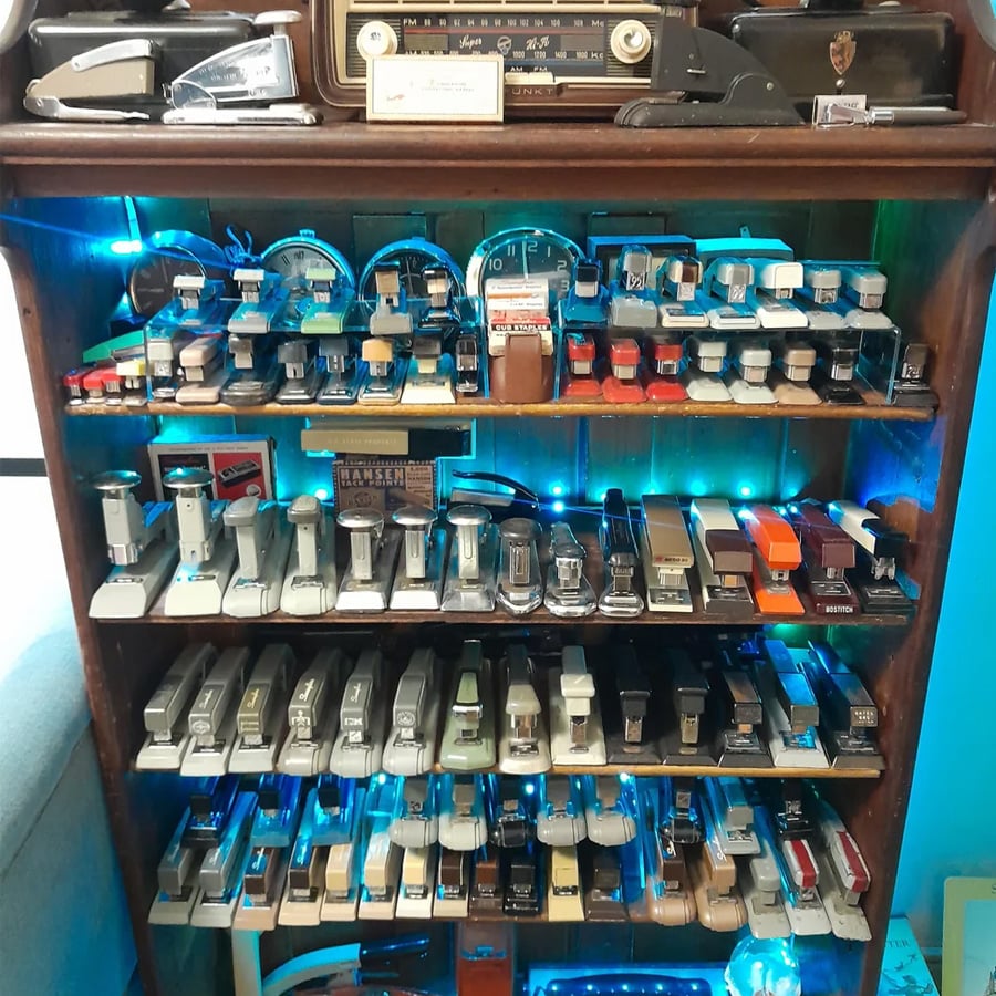 A wooden shelf displays an extensive collection of staplers arranged neatly in rows, with a vintage radio and tape dispensers on top and blue LED lights illuminating the shelves.