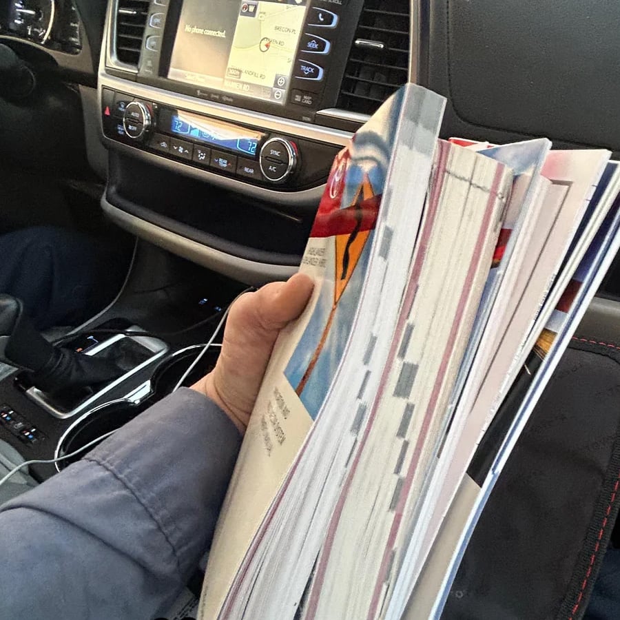 A person holding a thick stack of papers or magazines while sitting in the passenger seat of a car, with the dashboard, gear shift, and a connected phone visible.