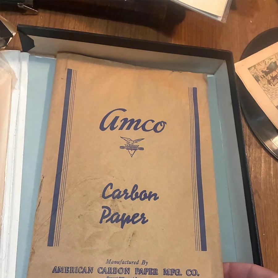 A vintage brown Amco Carbon Paper folder lies open in a box. The folder has blue text and lines, with “Manufactured by American Carbon Paper Mfg. Co.” printed at the bottom.