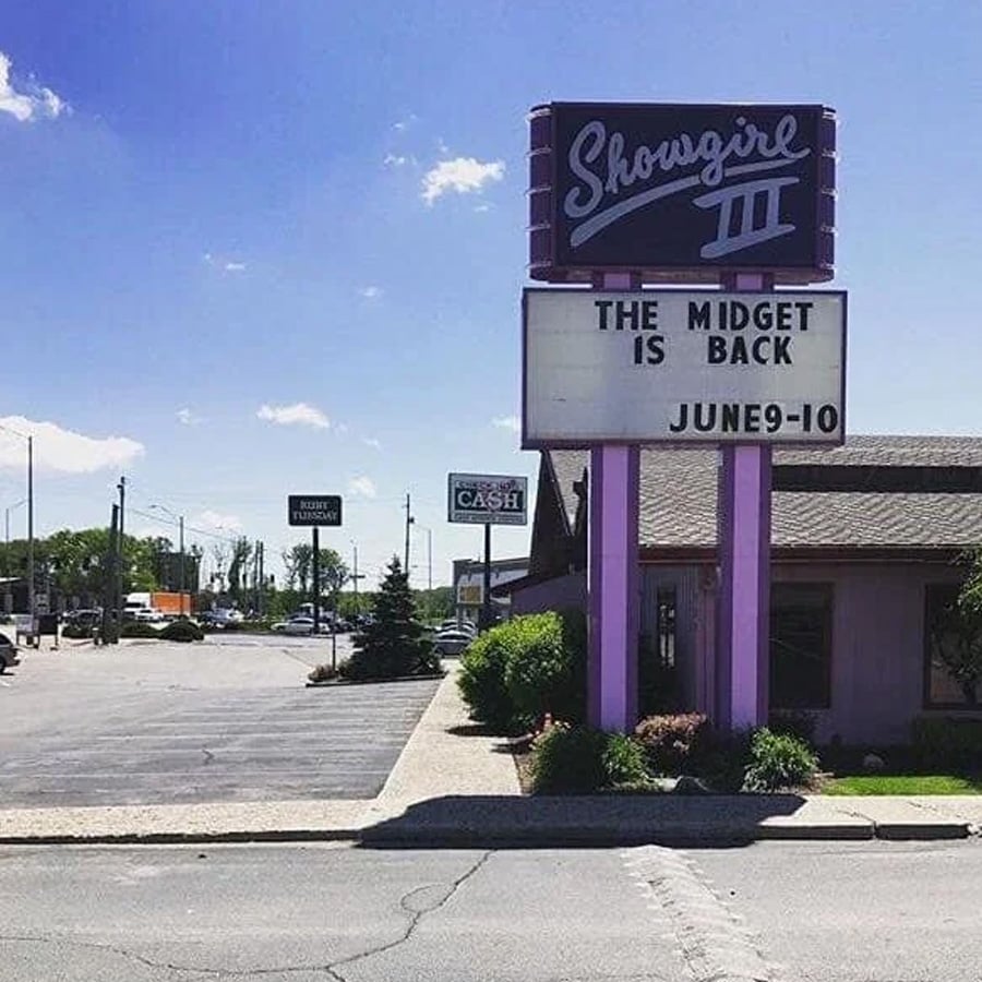 A roadside sign for “Showgirl III” reads “THE MIDGET IS BACK JUNE 9-10.” The building is purple, and the area has a parking lot and other businesses in the background.