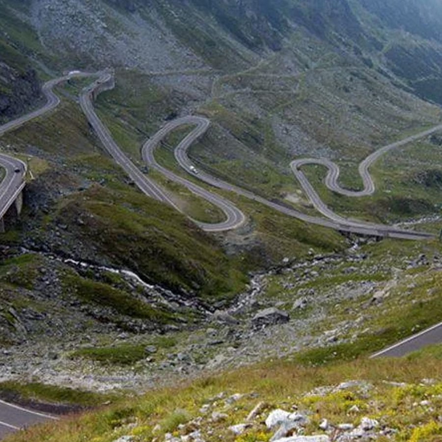A winding mountain road with many sharp curves snakes through green hills and rocky terrain, surrounded by steep slopes and sparse vegetation under a cloudy sky.