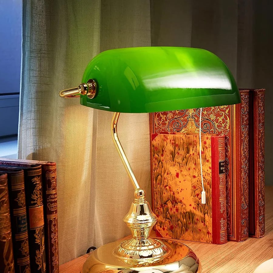 A classic green glass banker’s lamp with a brass base sits on a wooden desk, surrounded by ornate hardcover books, with a beige curtain in the background.