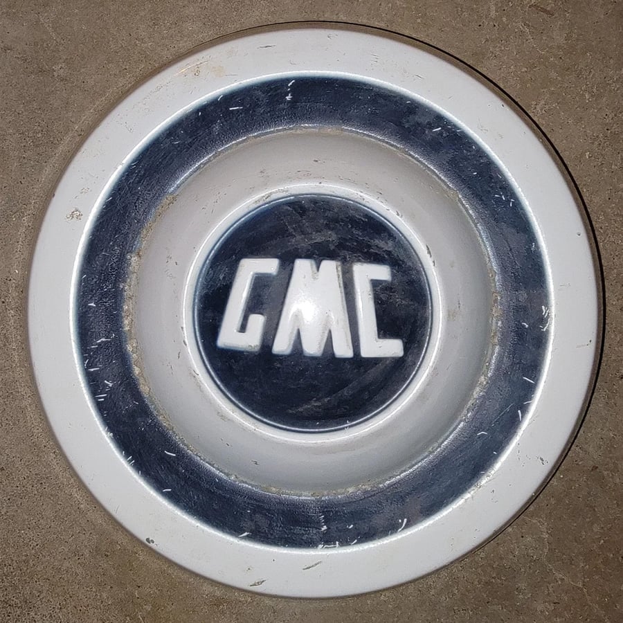 A close-up photo of a silver and white GMC car hubcap with the GMC logo in the center, sitting on a concrete surface.