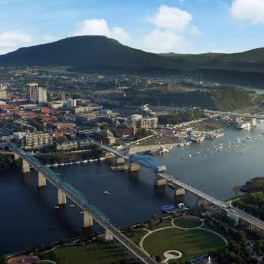 Aerial view of a city with bridges crossing a river, boats docked along the shore, downtown buildings, green parks, and a mountain in the background under a blue sky with scattered clouds.