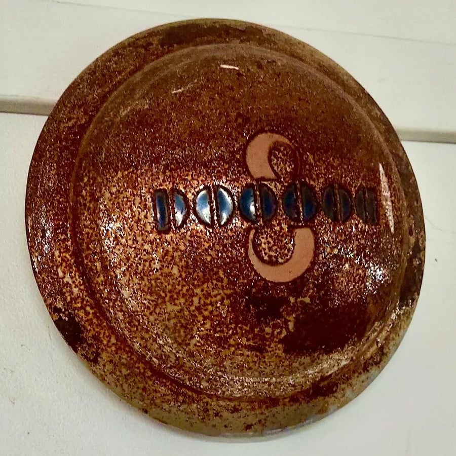 A round, brown, speckled ceramic wall plaque features a large tan “S” shape in the center with dark blue geometric accents across the middle.