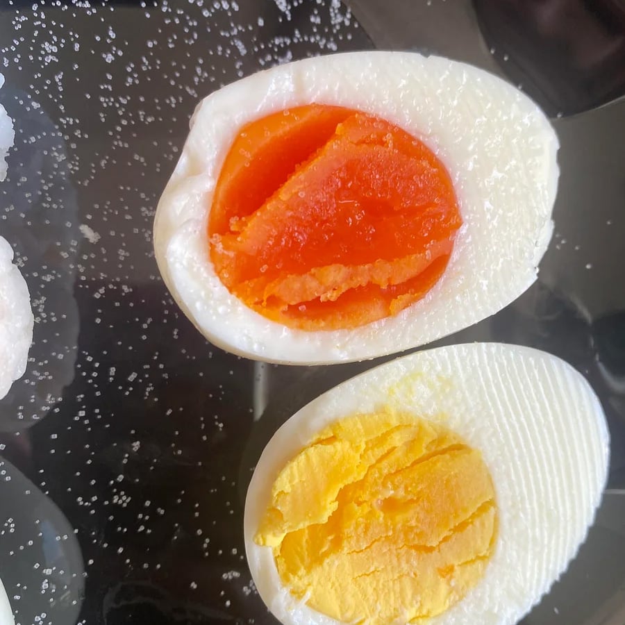 Two hard-boiled eggs cut in half on a black surface; one has a bright orange yolk, the other has a yellow yolk, both with scattered salt around them.