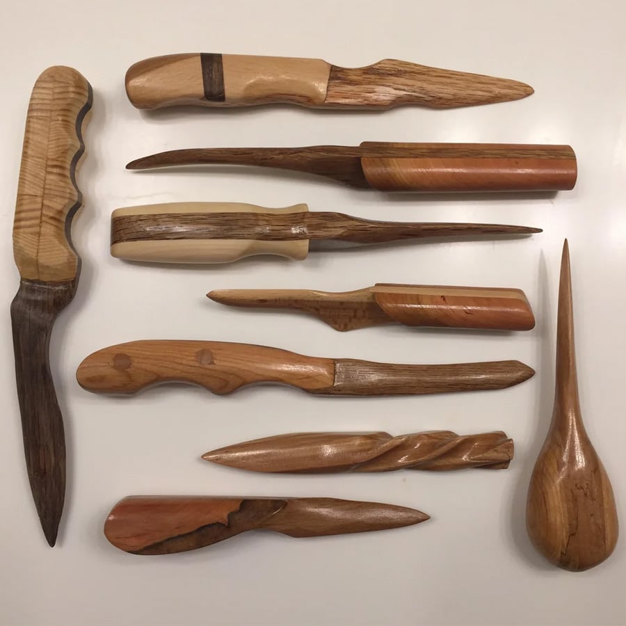 Ten handcrafted wooden letter openers in various shapes, sizes, and wood tones are arranged on a white surface. Each piece has a unique design, featuring curves, grooves, and smooth finishes.