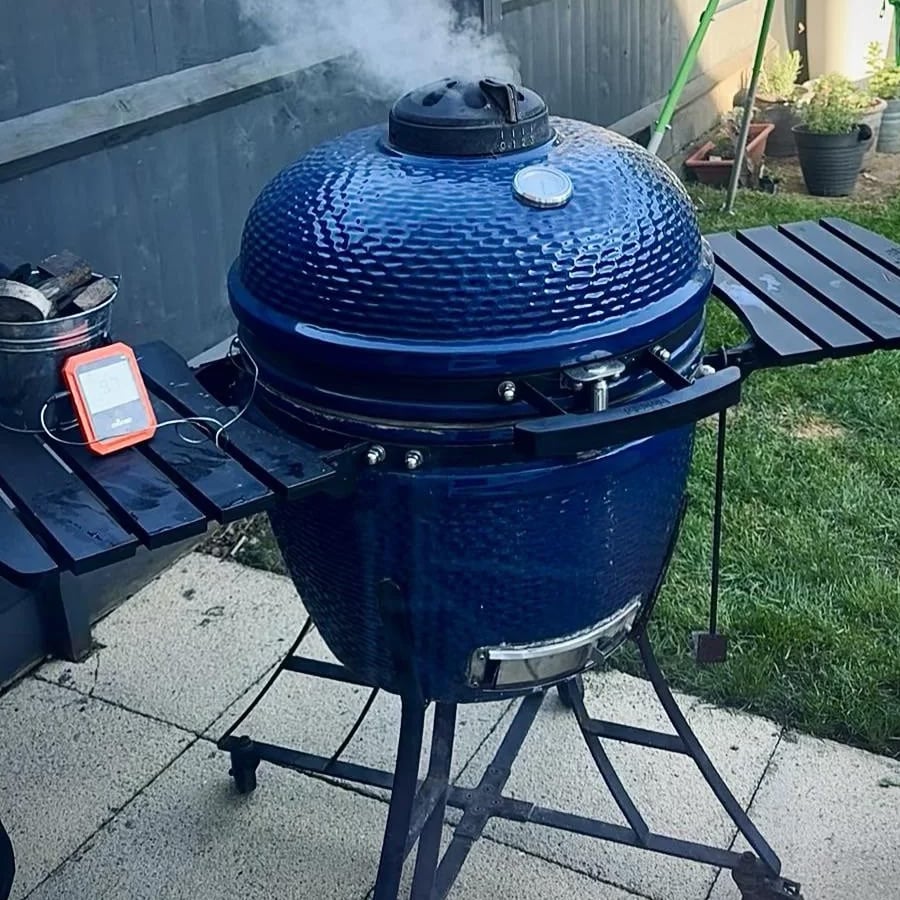 A blue ceramic grill with smoke coming from the top sits on a patio. A digital thermometer rests on a side shelf. Green grass and garden pots are visible in the background.