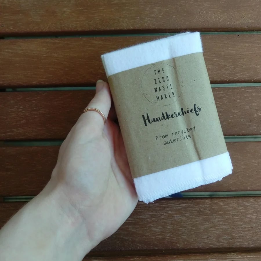 A hand holds a folded white handkerchief wrapped in brown paper labeled “The Zero Waste Maker Handkerchiefs From recycled materials” against a wooden background.