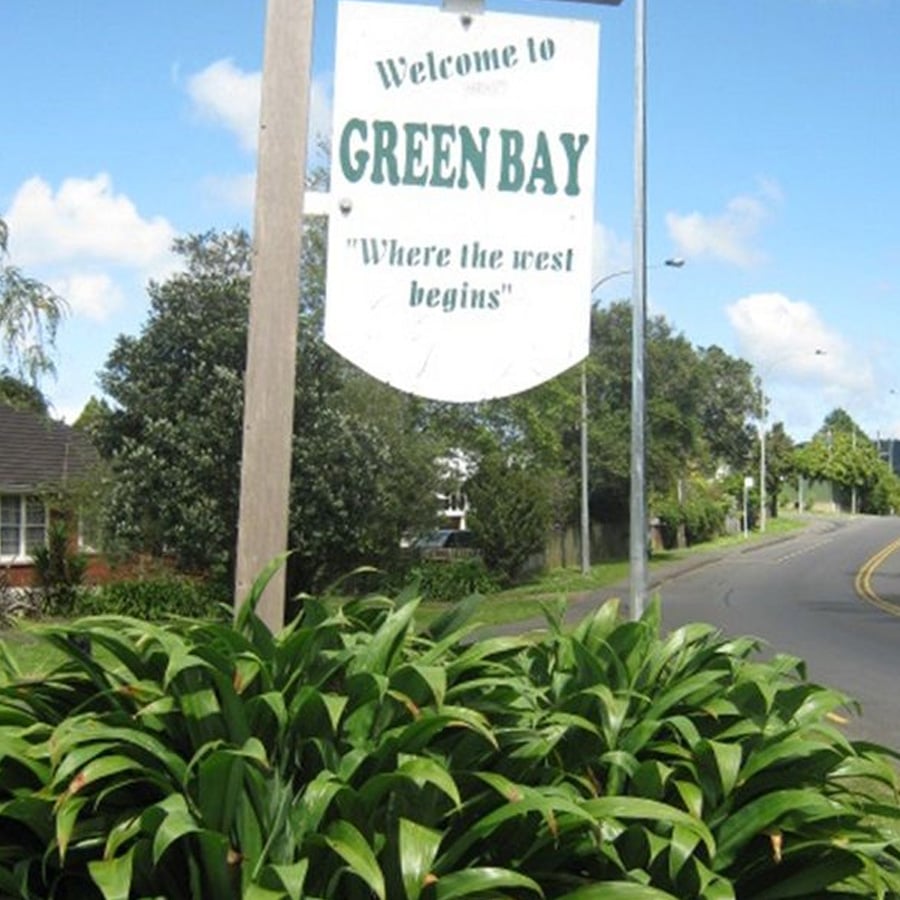 A street sign reads “Welcome to GREEN BAY ‘Where the west begins’” above green plants, with a road and houses in the background under a blue sky with clouds.