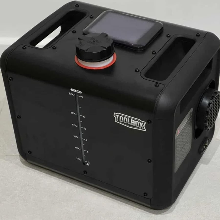 A black rectangular TOOLBOX device with a handle, a large cap on top, a vent on the side, and a vertical gauge marked with percentages. The device sits on a light-colored floor against a white wall.